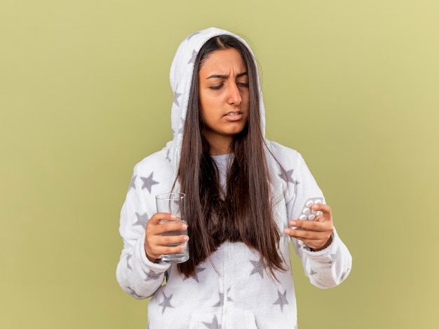 Thinking young ill girl putting on hood holding glass of water and looking at pills in her hand isolated on olive green background