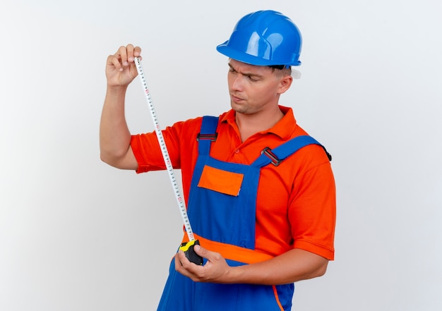 Foto gratuita giovane costruttore maschio di pensiero che indossa uniforme e casco di sicurezza che tiene e che esamina il nastro del tester su bianco