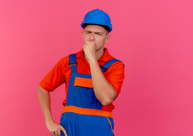 Thinking young male builder wearing uniform and safety helmet putting fist on chin on pink