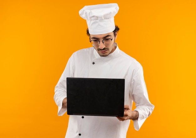Думающий молодой мужчина-повар в униформе шеф-повара и в очках держит и смотрит в ноутбук, изолированный на желтой стене