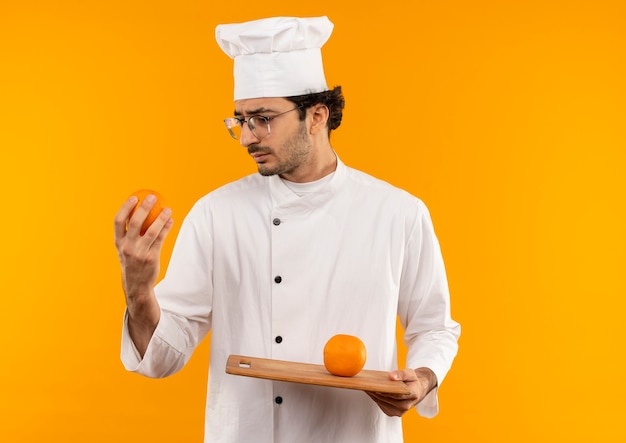 Pensando giovane cuoco maschio che indossa l'uniforme del cuoco unico e vetri che tengono e che esaminano arancio sul tagliere isolato sulla parete gialla