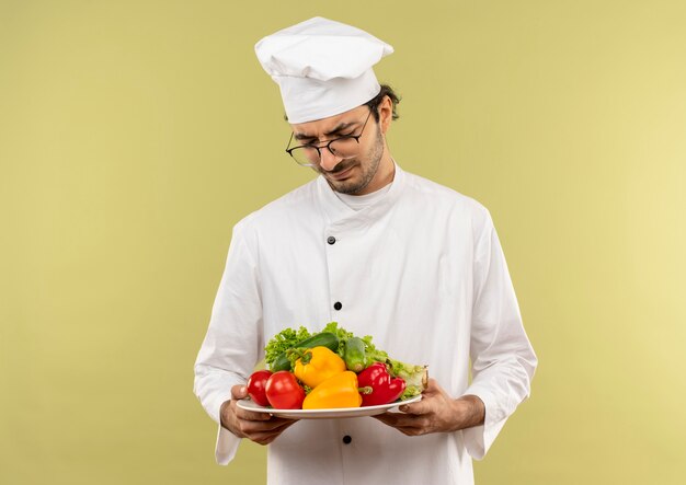 Думающий молодой мужчина-повар в униформе шеф-повара и в очках держит и смотрит овощи на тарелке, изолированной на зеленой стене
