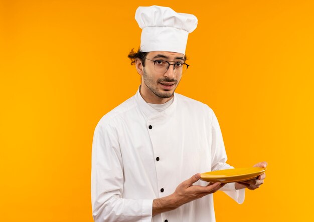 Thinking young male cook wearing chef uniform and glasses holding plate isolated on yellow wall