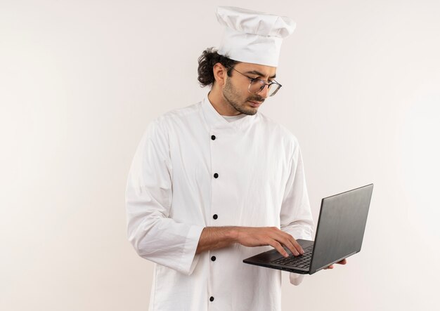 Thinking young male cook wearing chef uniform and glasses holding and used laptop isolated on white wall