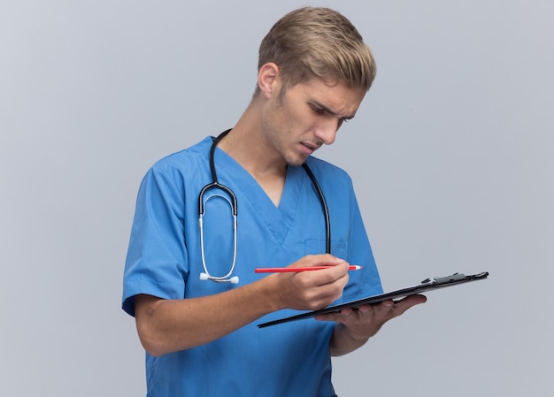Thinking young male doctor wearing doctor uniform with stethoscope writing something on clipboard isolated on white wall