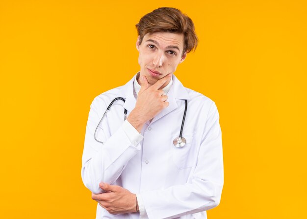 Thinking young male doctor wearing medical robe with stethoscope grabbed chin isolated on orange wall