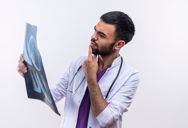 Thinking young male doctor wearing stethoscope medical gown looking at x-ray on his hand on isolated white