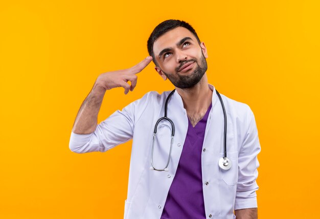 Thinking young male doctor wearing stethoscope medical gown put his finger on forehead on isolated yellow wall