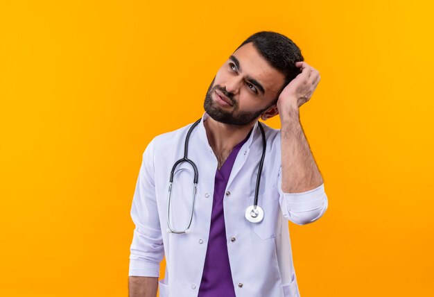 Думающий молодой мужчина-врач в медицинском халате со стетоскопом почесывает голову на изолированной желтой стене