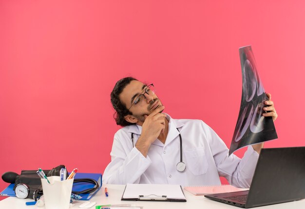 Thinking young male doctor with medical glasses wearing medical robe with stethoscope sitting at desk