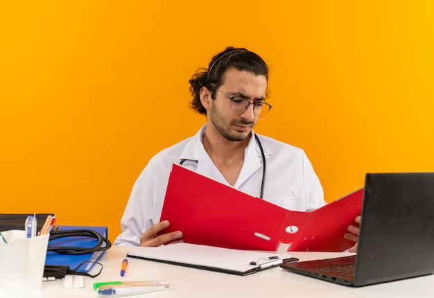 Thinking young male doctor with medical glasses wearing medical robe with stethoscope sitting at desk