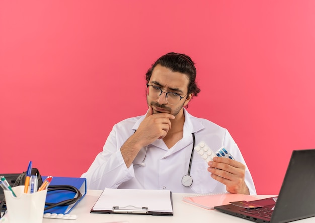 Thinking young male doctor with medical glasses wearing medical robe with stethoscope sitting at desk