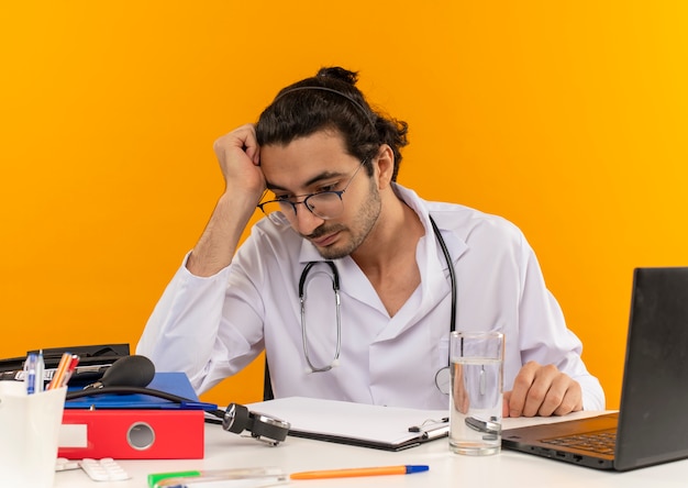 Free photo thinking young male doctor with medical glasses wearing medical robe with stethoscope sitting at desk