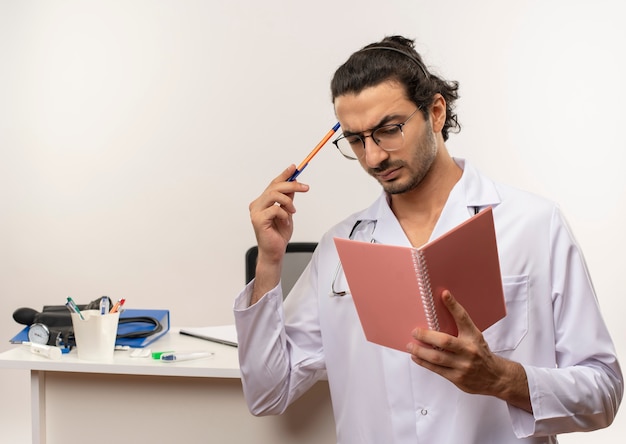 Foto gratuita pensando giovane medico maschio con occhiali medici che indossano accappatoio medico con stetoscopio in piedi davanti alla scrivania