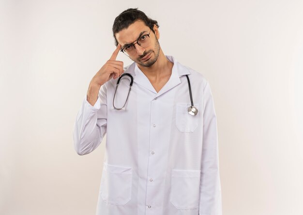 Thinking young male doctor with optical glasses wearing white robe with stethoscope putting finger to forehead on isolated white wall with copy space