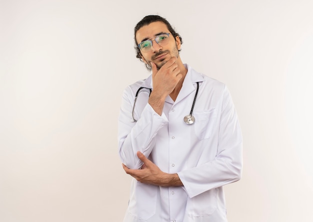 Thinking young male doctor with optical glasses wearing white robe with stethoscope putting hand on chin on isolated white wall with copy space