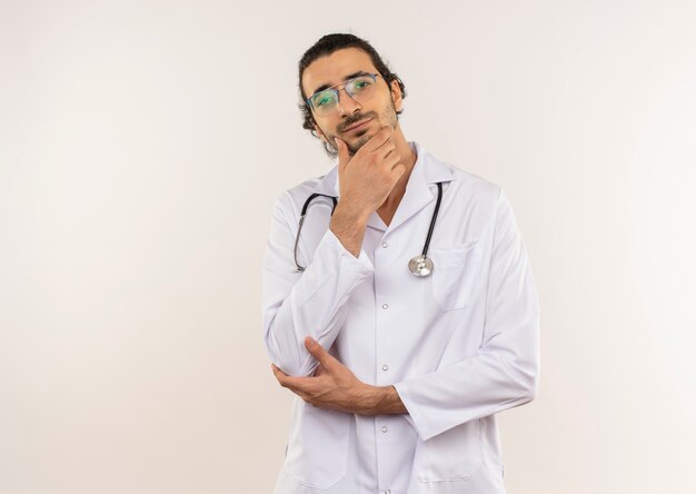 Thinking young male doctor with optical glasses wearing white robe with stethoscope putting hand on chin on isolated white wall with copy space