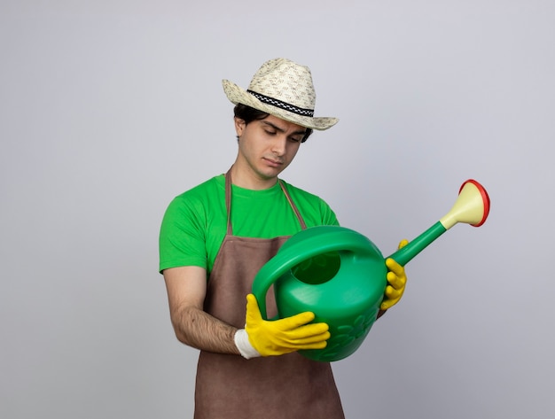 Pensando giovane giardiniere maschio in uniforme che indossa guanti e cappello da giardinaggio tenendo e guardando annaffiatoio