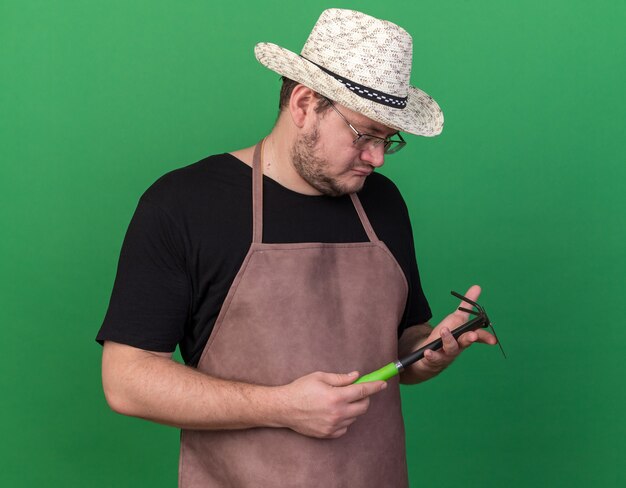 Thinking young male gardener wearing gardening hat holding and looking at hoe rake isolated on green wall