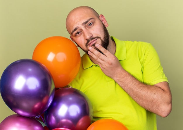 Thinking young man wearing yellow t-shirt holding balloons grabbed chin isolated on olive green wall