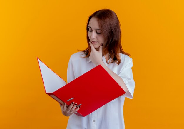 黄色で隔離されたフォルダーとつかんだあごを持って見て若い赤毛の女の子を考えて