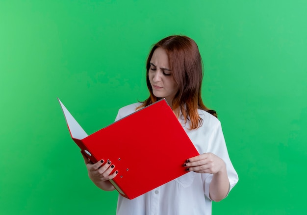 Думающая молодая рыжая девушка держит и смотрит на папку, изолированную на зеленом