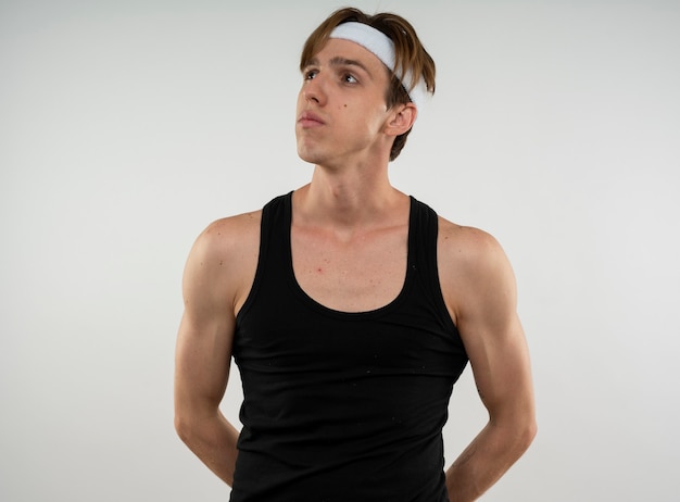 Thinking young sporty guy looking at side wearing headband and wristband holding hands on waist isolated on white wall