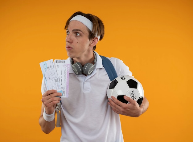 Thinking young sporty guy wearing headband with wristband and backpack with headphones on neck holding tickets with ball isolated on yellow wall