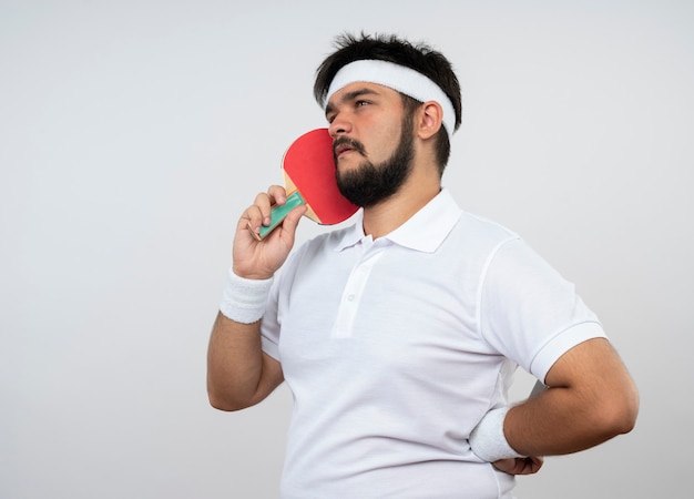 Думающий молодой спортивный человек смотрит в сторону в головной повязке и браслете, держа ракетку для пинг-понга на щеке, положив руку на бедро, изолированную на белой стене