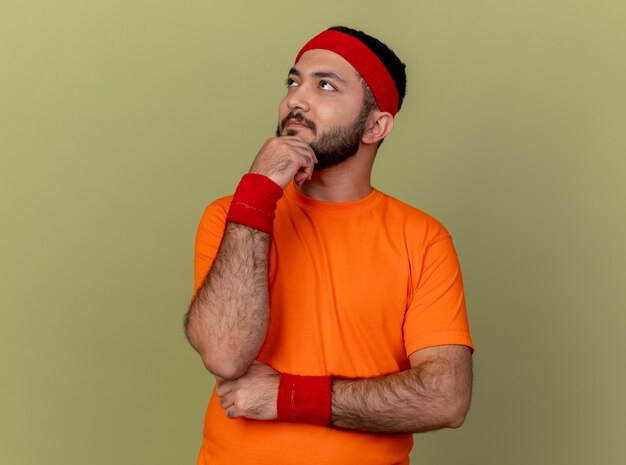 Thinking young sporty man looking at side wearing headband and wristband putting hand on chin isolated on olive green background