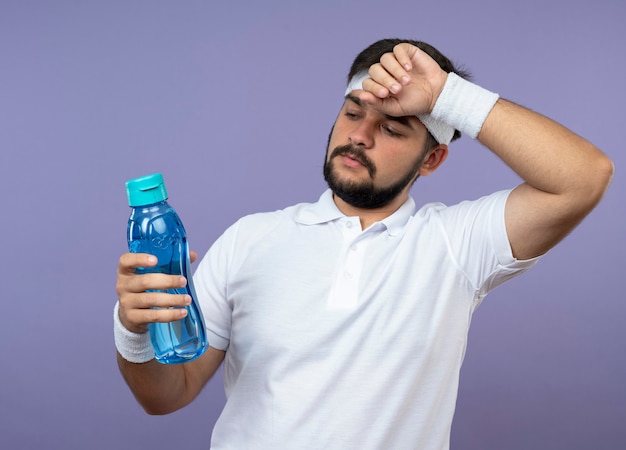 Pensando giovane uomo sportivo che indossa fascia e polsino tenendo e guardando la bottiglia d'acqua mettendo la mano sulla fronte isolata sulla parete verde