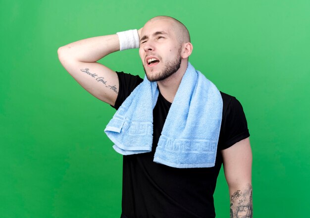 Thinking young sporty man wearing wristband putting hand on behind head with towel on shoulder isolated on green wall