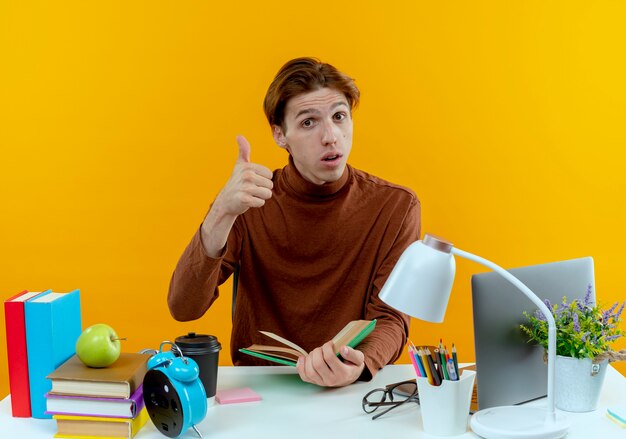Thinking young student boy sitting at desk with school tools holding book his thumb up on yellow