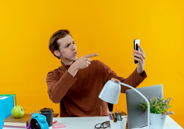 Thinking young student boy sitting at desk with school tools take a selfie and showing you gesture on yellow