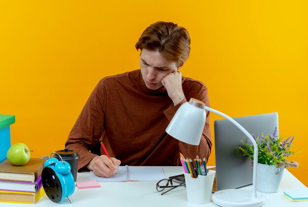 黄色のノートに何かを書いている学校の道具を持って机に座っている若い学生の少年を考える