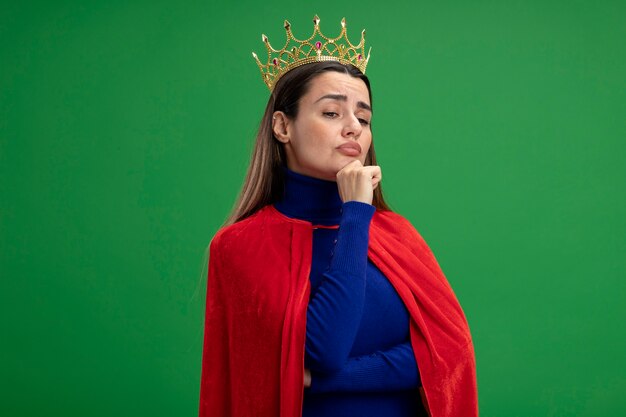 Foto gratuita pensando la giovane ragazza del supereroe guardando al lato che indossa la corona ha afferrato il mento isolato su sfondo verde