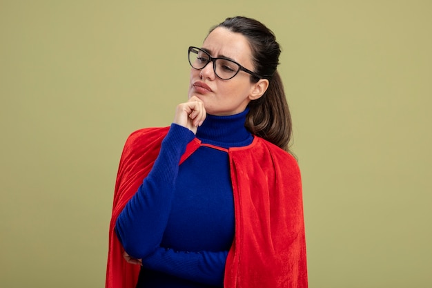 Thinking young superhero girl looking at side wearing glasses putting hand on chin isolated on olive green background