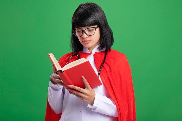 Thinking young superhero girl wearing stethoscope with medical robe and cloak with glasses holding and looking at book isolated on green wall