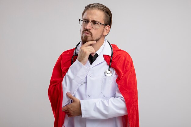 Thinking young superhero guy looking at side wearing medical robe with stethoscope and glasses grabbed chin isolated on white background