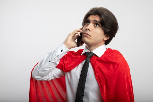 Думающий молодой парень супергероя смотрит вверх в галстуке, разговаривает по телефону, изолированном на белом фоне