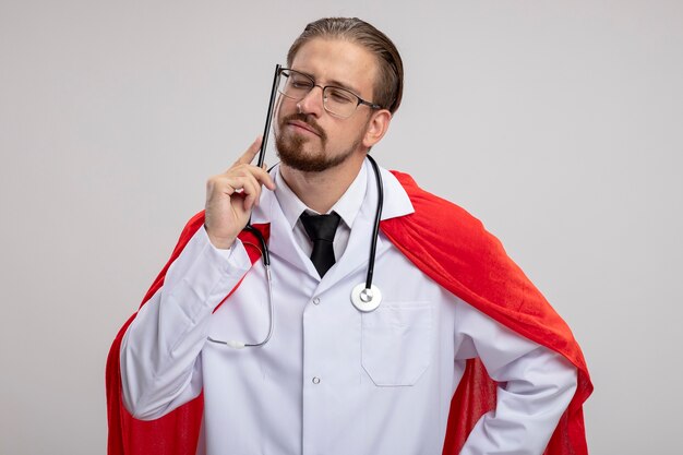 Thinking young superhero guy wearing medical robe with stethoscope and glasses putting pencil on cheek isolated on white background