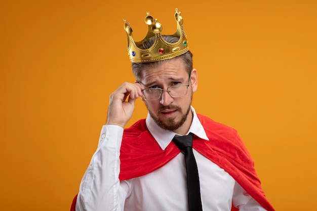 Thinking young superhero guy wearing tie and crown wearing and grabbed glasses isolated on orange background