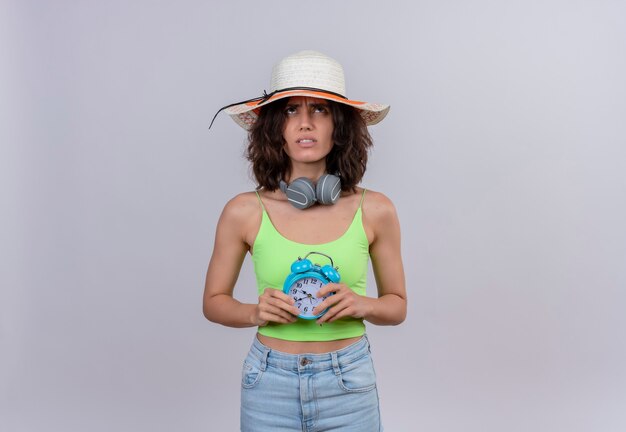 A thinking young woman with short hair in green crop top wearing sun hat looking up holding blue alarm clock on a white background