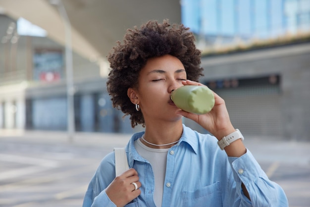 Thirsty curly haired young woman drinks fresh water from bottle wears blue shirt carries bag keeps eyes closed strolls outdoors poses against blurred background Hydration and aqua balance regulation