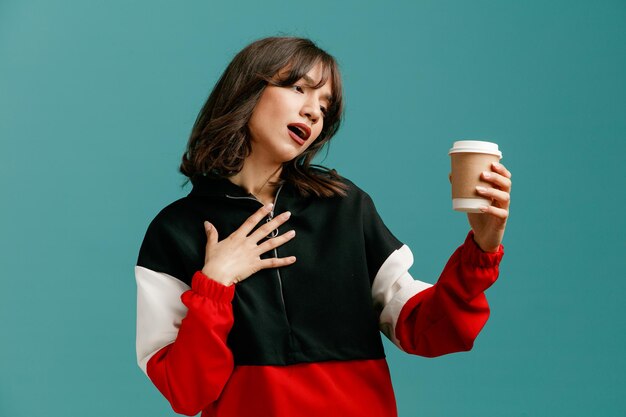 Thirsty young caucasian woman holding and looking at paper coffee cup while keeping hand on chest isolated on blue background