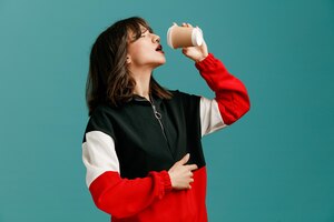 thirsty young caucasian woman standing in profile view holding paper coffee cup near mouth while keeping hand on belly with closed eyes isolated on blue background