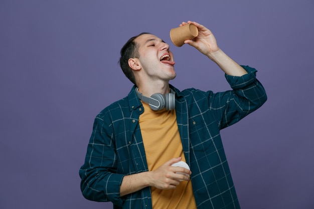 口の上に紙のコーヒー カップと紫色の背景に分離された舌を示す口を開けてカップに別の手でそのキャップを保持している首の周りにヘッドフォンを身に着けているのどが渇いて若い男子学生