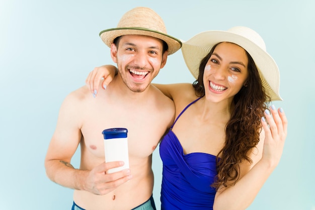 Это лучший солнцезащитный лосьон. Портрет веселой молодой пары с солнцезащитным кремом на лице, улыбающейся и держащей бутылку солнцезащитного крема