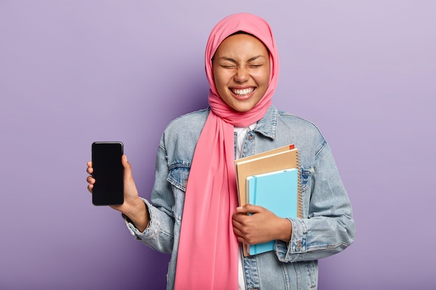 This is phone you need. Joyful woman with Islamic views, wears traditional hijab, shows smartphone screen and laughs