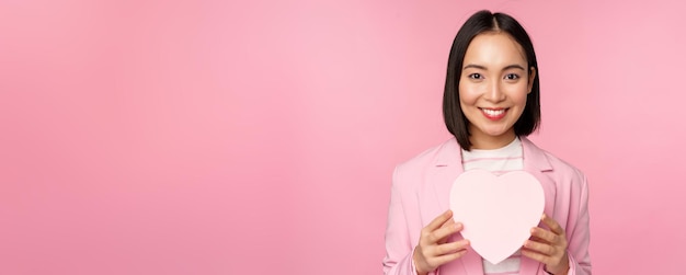 This is for you Romantic cute asian corporate woman girl in suit showing heartshaped box with gift standing over pink background
